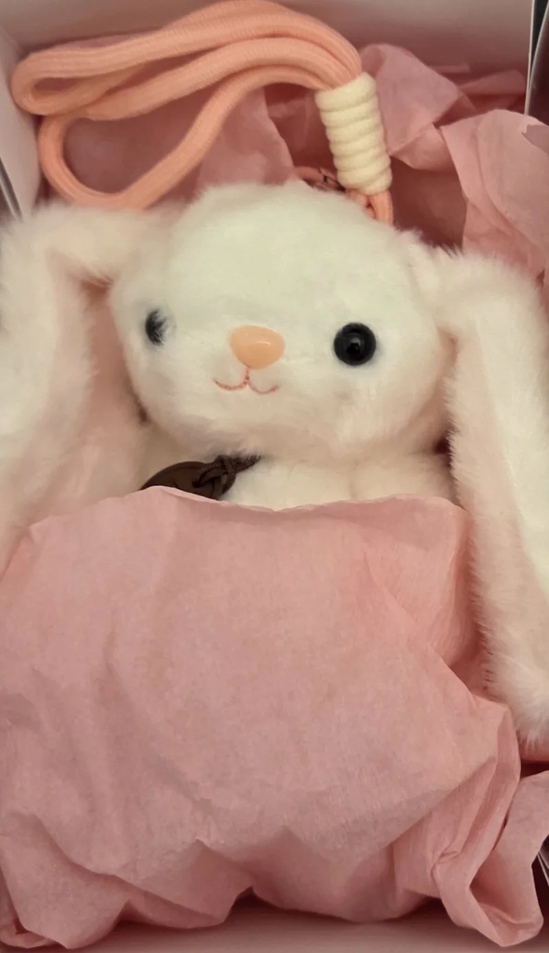Un conejito blanco de orejas largas asomándose entre papel crepe rosa dentro de su cajita